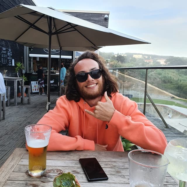 Profile pic of Ulysse Coates in a salmon coloured jumper, doing the surf sign with his first and last fingers